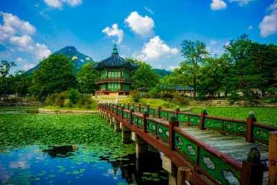 Looking at the beautiful Hyangwonjeong Pavilion sitting on a small island in the middle of Hyangwonji pond inside Gyeongbok Palace grounds. Bright green leaves on the trees and clear blue skies can be seen surrounding the building